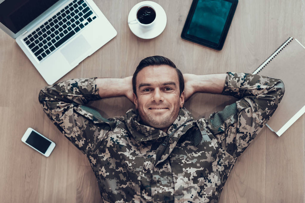 Military Man Lying on Table With Modern Gadgets. Cheerful Smiling Handsome Brown Haired Person in Military Uniform Surrounded by Laptop, Tablet, Smartphone and Cup of Coffee - 写真・画像