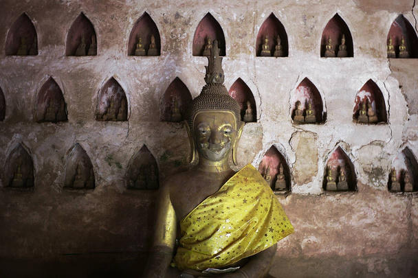 Vientiane / Laos - JUL 06 2011: estatuas budistas teravadas bañadas en oro en un monasterio con un paño de seda amarillo
 - Foto, imagen