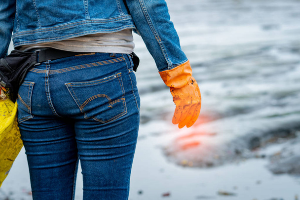 Εθελοντές φορούν τζιν και πουκάμισο με μακριά μανίκια και φορούν πορτοκαλί καουτσούκ γάντια για να συλλέγουν τα σκουπίδια στην παραλία. Περιβάλλον παραλία. Γυναίκα καθαρισμός στην παραλία. Συγύρισμα σκουπίδια στην παραλία. - Φωτογραφία, εικόνα