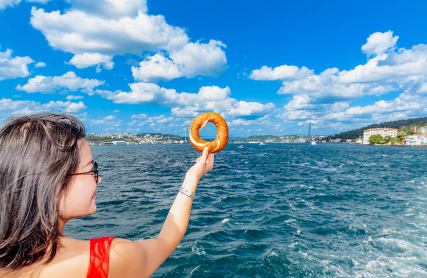 Beautiful Chinese woman holds traditional Turkish simit (bagel) with view of Bosphorus in Istanbul,Turkey - Foto, afbeelding