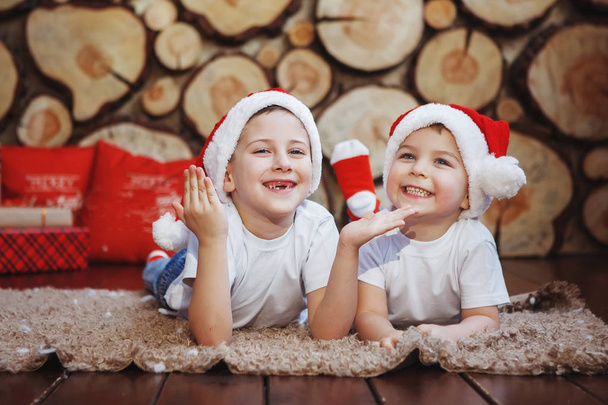 Dwóch chłopców brat w Swiąteczne leżącego i śmiejąc się na dywanie w pobliżu drzewa nowy rok w tle kawałki drzewa. - Zdjęcie, obraz