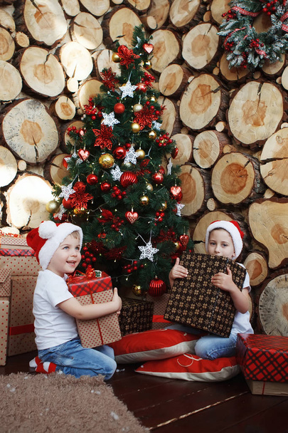 Zwei Brüder in Weihnachtsmützen sitzen mit Geschenkschachteln in der Hand auf einem Karo neben dem Neujahrsbaum im Hintergrund der Baumfällungen. - Foto, Bild
