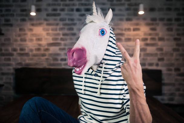 Freaky young man in comical mask sits on bed in stylish apartment. Funny unicorn - Foto, Imagen
