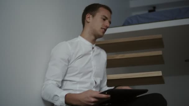 Side view of a young professional man freelancer in white shirt using a laptop computer while sitting down on modern staircase. Low angle footage - Video