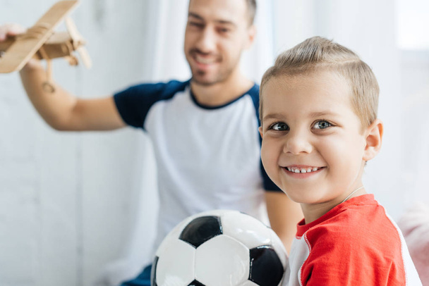 selective focus of smiling boy with football ball and father with wooden toy plane at home - Fotoğraf, Görsel