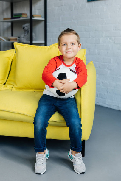 niño sonriente con pelota de fútbol en las manos descansando en el sofá amarillo en casa
 - Foto, Imagen