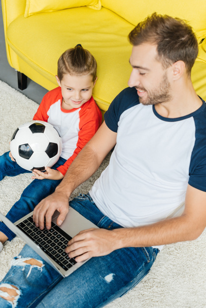 man using laptop while little son with football ball sitting near by on floor at home - Valokuva, kuva