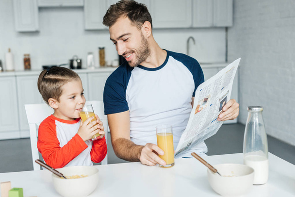 father with newspaper looking at son drinking juice during breakfast in kitchen - Photo, Image