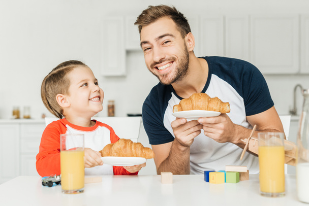 Lächelnder junger Vater hält Croissant in der Hand und blickt in die Kamera, während er mit Sohn zu Hause frühstückt - Foto, Bild