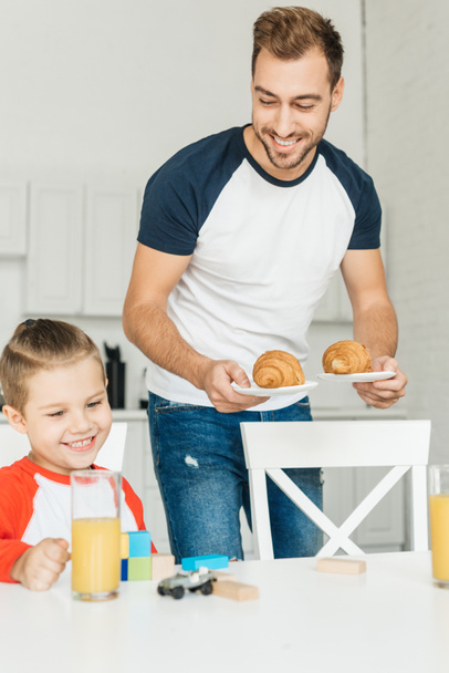 handsome young father serving croissants for breakfast with son while he playing with toys - Fotó, kép