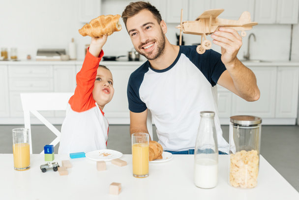 handsome young father and son playing with croissant and toy plane while having breakfast together at home - Фото, изображение
