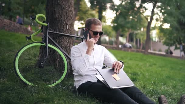 Young successful businessman in white shirt and black glasses. Man sit on grass ground, work on laptop and talks on mobile in city park on green lawn outdoors on nature leans on his bike neat the tree - Footage, Video