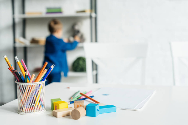 color pencils and wooden blocks on table with album lying on table with little kid blurred on background - Foto, Bild