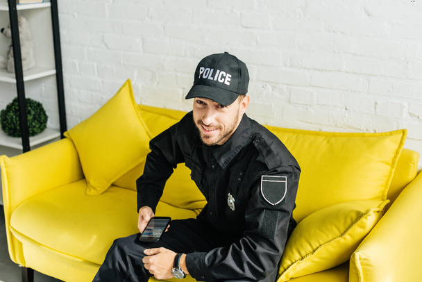 happy young policeman with smartphone sitting on yellow couch and looking at camera - Фото, зображення