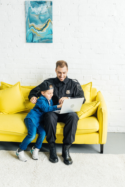 handsome young father in police uniform and son using laptop together while sitting on yellow couch at home - Photo, Image