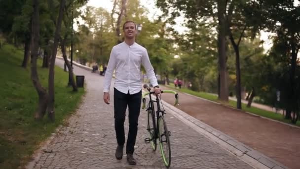 Full length of caucasian smiling young man in white shirt walking with bicycle on the street in town. Rolling his trekking bike while walking by park. Front view - Video