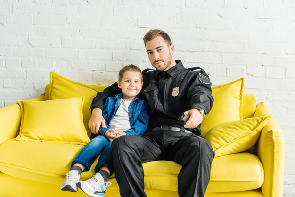 mladý otec v policejní uniformě sledování televize se synem, zatímco sedí na žluté pohovce doma - Fotografie, Obrázek