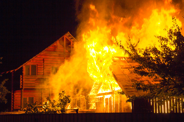 夜は、木造住宅を燃焼します。明るいオレンジ色の炎と暗い空、木のシルエットと住宅隣人コテージ背景に瓦屋根の下から濃い煙。災害や危険の概念. - 写真・画像