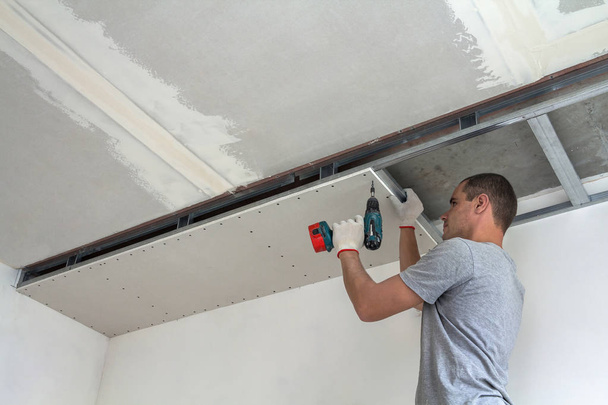 Young man in usual clothing and work gloves fixing drywall suspended ceiling to metal frame using electrical screwdriver on ceiling insulated with shiny aluminum foil. DIY, do it yourself concept. - Photo, Image