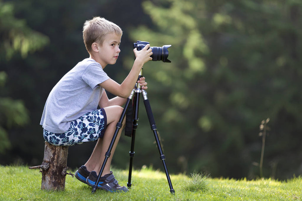 Jeune garçon blond assis sur un tronc d'arbre sur une clairière herbeuse prenant des photos avec une caméra trépied
. - Photo, image