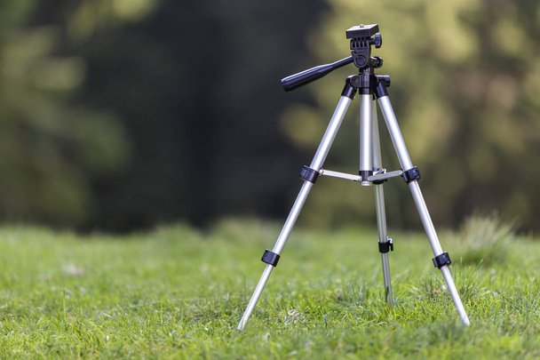 Isolated aluminum shiny tripod standing alone in fresh grassy meadow on bright sunny summer day on blurred dark green forest trees foliage bokeh copy space background. Photography as job and hobby. - Fotoğraf, Görsel