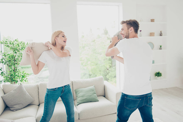 Beautiful attractive adorable lovely charming cute nice couple in casual white t-shirts and jeans, fighting with pillows, light interior - Photo, Image