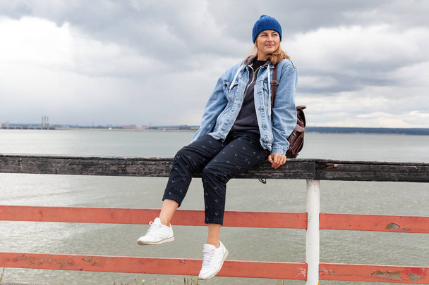 Retrato de una hermosa modelo joven en azul sombrero de punto sentarse cerca del mar. Fotografía cálida de otoño.Mujer sonriendo y mirando hacia otro lado, alegre humor alegre
. - Foto, imagen