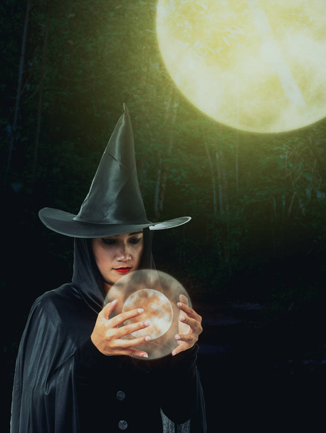 Πορτραίτο γυναίκας στο μαύρο Scary μάγισσα κοστούμι αποκριών κρατώντας κρυστάλλινη μπάλα με φως του φεγγαριού σε ένα σκοτεινό δάσος - Φωτογραφία, εικόνα