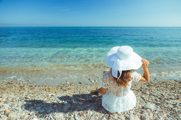 The back view of young beautiful woman in white dress sitting on the beach and looking at the sea horizon, image with copy space - concept of traveling and luxury vacation In Italy - shot from behind - Fotografie, Obrázek