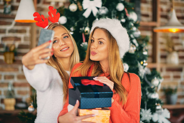 Girlfriends taking selfie and holding gifts while standing in front of Christmas tree. Christmas and winter holidays concept. - Photo, Image