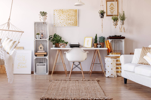 White chair placed by the desk with fresh plants and typewriter in real photo of bright living room interior with posters and rug on the floor - Zdjęcie, obraz