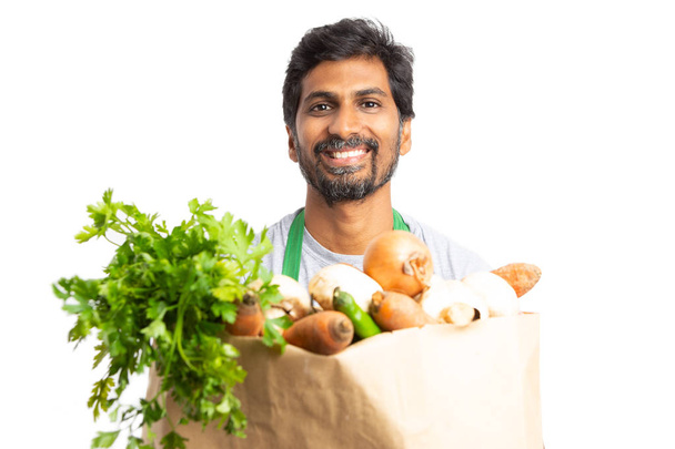 Uśmiechnięty mężczyzna pracownika indyjskich w sklepie lub rynku trzymając brązową papierową torbę z świeże produkty spożywcze na białym tle - Zdjęcie, obraz