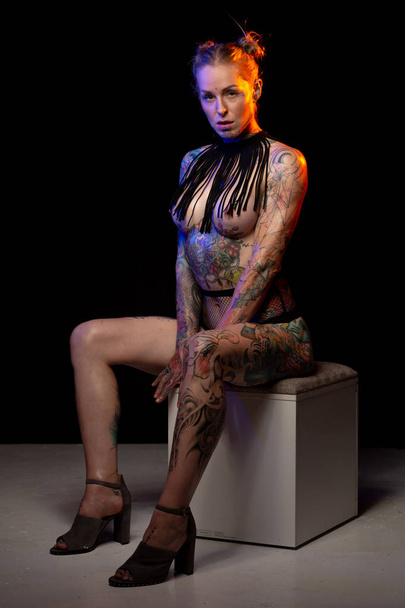 Muscled caucasian woman with tattoos and oiled skin wearing heels poses under colored lights on stool - Valokuva, kuva