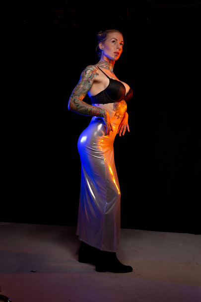 Mulher branca musculada com tatuagens e pele oleada vestindo poses saia brilhante sob luzes coloridas
 - Foto, Imagem