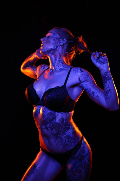 Musculosa mujer caucásica con tatuajes y piel aceitada posa bajo luces de colores
 - Foto, imagen
