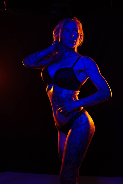 Muscled caucasian woman with tattoos and oiled skin poses under colored lights - Photo, image