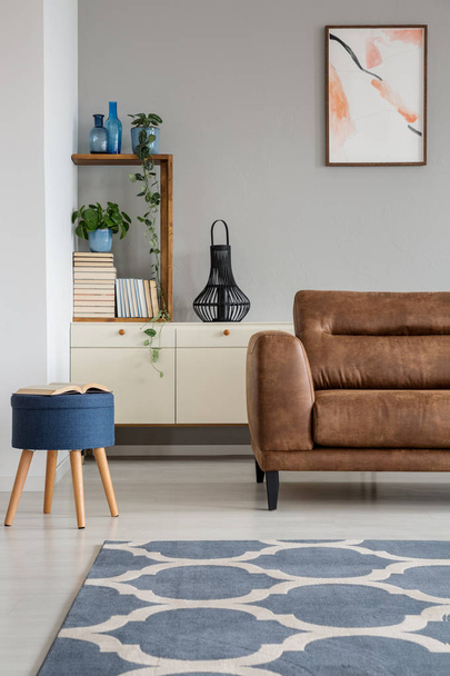 Navy blue stool next to leather sofa in grey living room interior with poster and carpet. Real photo - Φωτογραφία, εικόνα