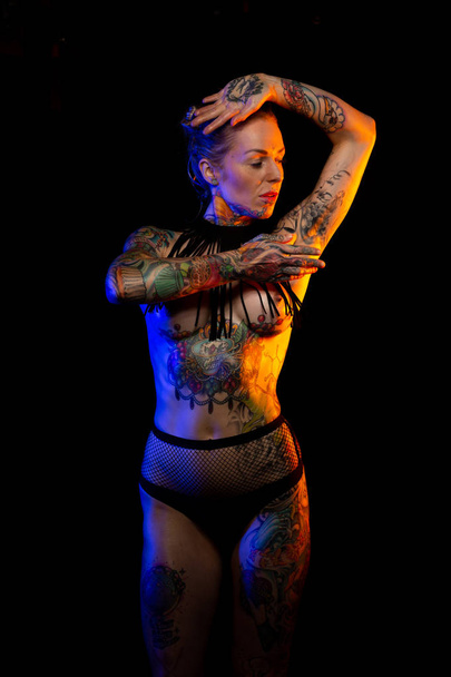 Muscled caucasian woman with tattoos and oiled skin wearing necklace and fishnet underwear poses under colored lights - 写真・画像