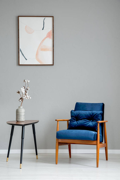 Flowers on wooden table next to blue armchair in grey apartment interior with poster. Real photo - Photo, Image