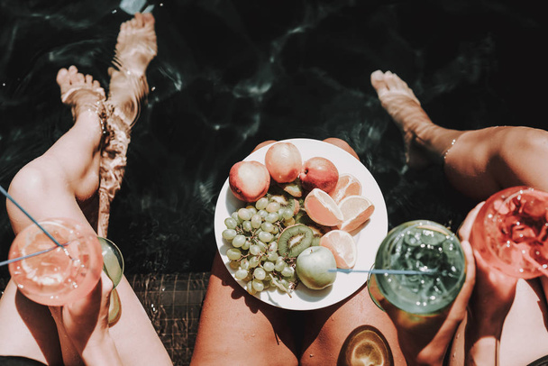 Young Women with Fruits and Cocktails at Poolside. Closeup of Group of Young Beautiful Women sitting at Poolside holding Colorful Alcoholic Cocktails and Bowl of Fresh Fruits. Summer Vacation Concept - Foto, immagini