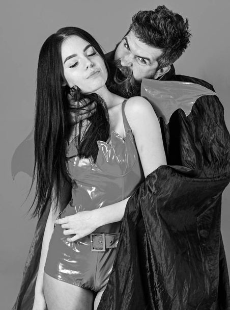 Vampire bites female neck. Couple in love play role game. Vampires victim concept. Man and woman dressed like vampire, demon, red background. Vampire in cloak behind sexy devil girl - Фото, зображення