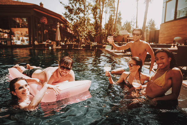 Group of Young Happy People Swimming in Pool. Young Smiling Friends wearing Sunglasses Laughing and Relaxing Together in Outdoor Hotel Pool next to Poolside. Summer Vacation Concept. Pool Party - 写真・画像