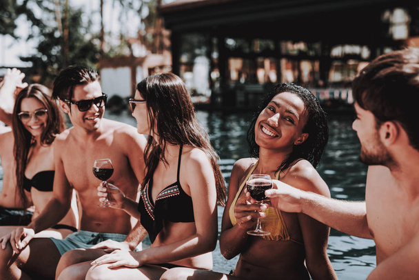 Young Smiling Friends Drinking Wine at Poolside. Group of Young Happy People Sitting at Poolside Together Talking and holding Glasses of Wine. Happy Friends Enoying Pool Party. Summer Vacation Concept - Photo, Image