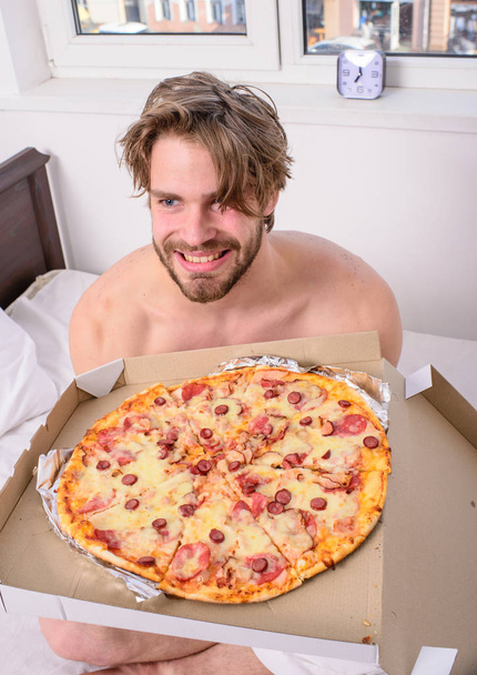 Food delivery service. Man bearded handsome bachelor eating cheesy food for breakfast in bed. Man likes pizza for breakfast. Break diet concept. Guy holds pizza box sit bed in bedroom or hotel room - Foto, Bild