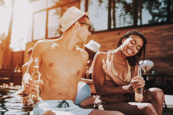 Smiling Couple with Alcoholic Drinks at Poolside. Beautiful Black Woman and Young Man holding Bottles of Beer and having Fun at Poolside. Happy Friends Enoying Pool Party. Summer Vacation Concept - Фото, изображение