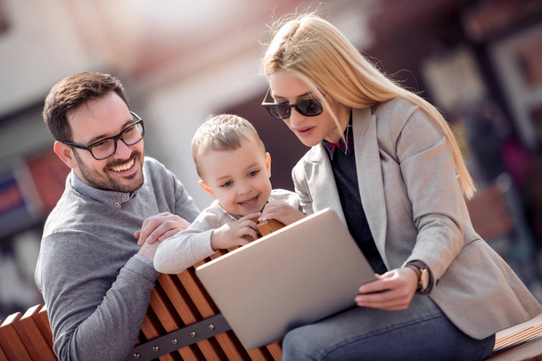 Jonge ouders en klein kind met behulp van laptop zittend in stadspark - Foto, afbeelding
