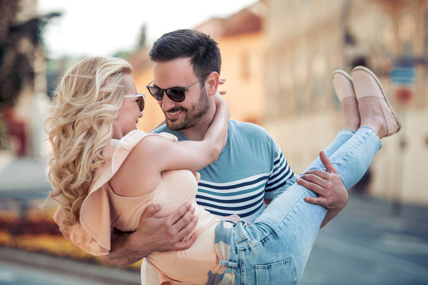sorridente jovem segurando namorada em braços ao ar livre à luz do dia
 - Foto, Imagem