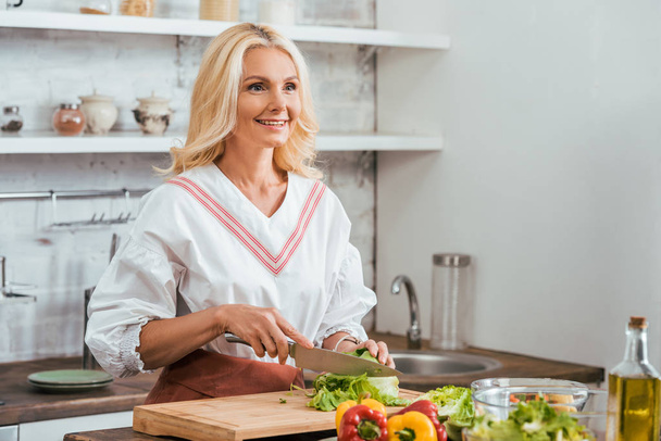 smiling attractive adult woman preparing salad for dinner, cutting cabbage and looking away at home - Foto, immagini