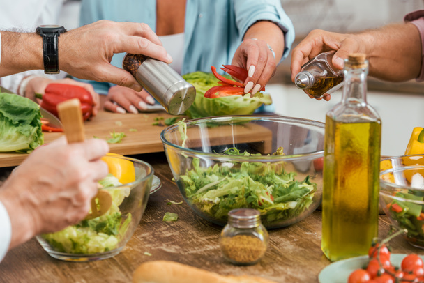 cropped image of mature friends preparing salad for dinner at home and adding spices in bowl - Photo, Image