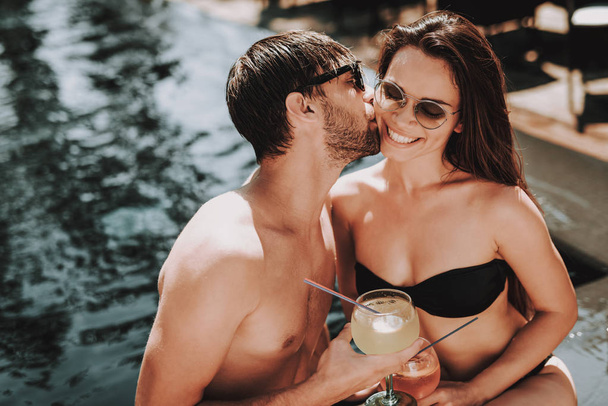 Smiling Couple Drinking Cocktails at Poolside. Beautiful Young Couple holding Glasses with Alcoholic Drinks and Kissing at Poolside. Happy Friends Enoying Pool Party. Summer Vacation Concept - Foto, Imagem